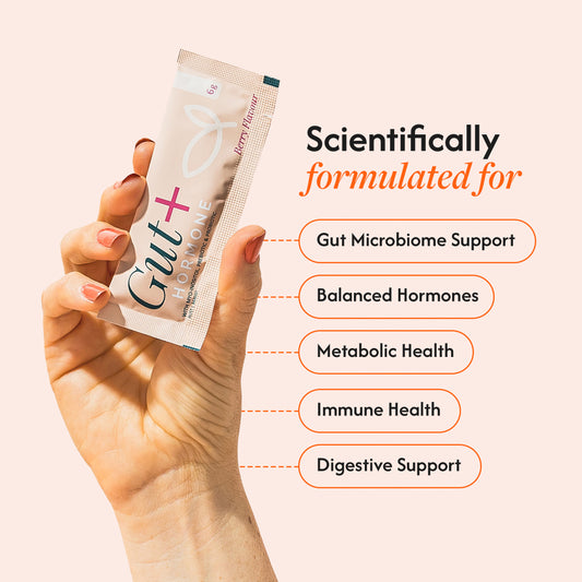 A hand holds a single packet of Gut+ Hormone supplement powder against a pale peach background, with marketing text listing benefits: Gut Microbiome Support, Balanced Hormones, Metabolic Health, Immune Health, and Digestive Support.